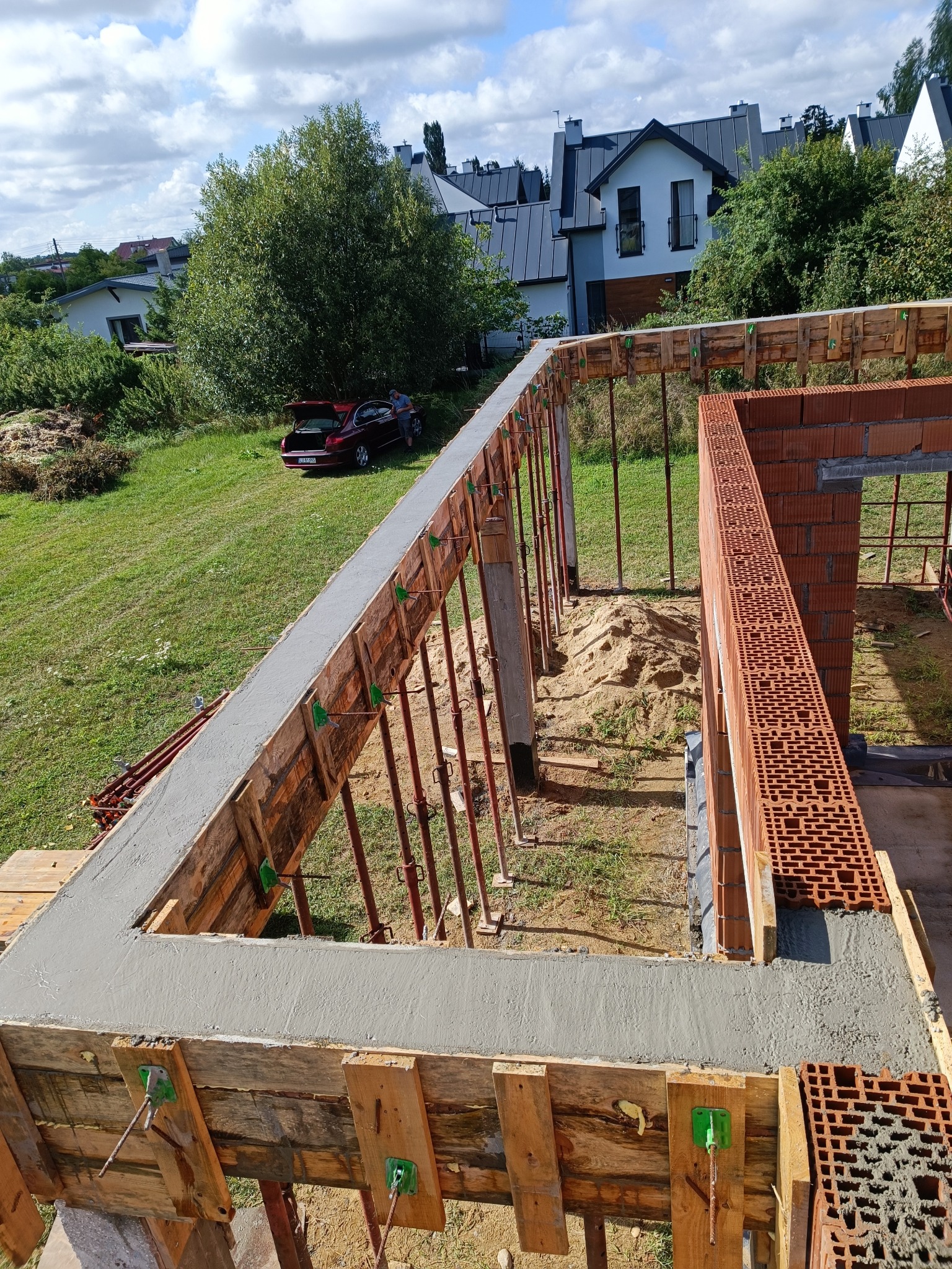 Wylany betonowy wieniec w szalunku, widoczne podpory i fragment muru z czerwonej cegły, w tle domy i samochód osobowy