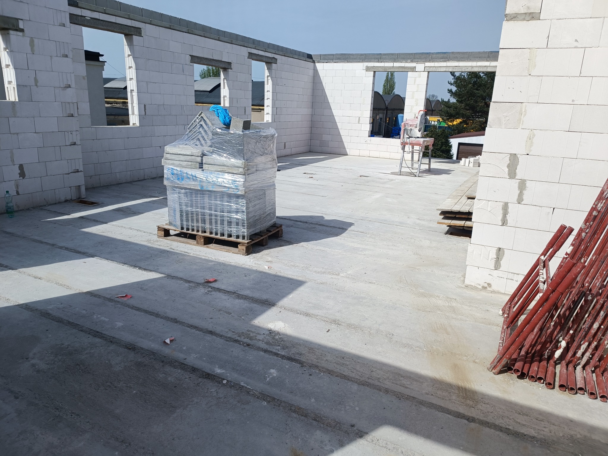 Wnętrze surowego domu murowanego w trakcie budowy, widoczne ściany z białych bloczków, otwory okienne, paleta z materiałami budowlanymi i czerwone rusztowania w rogu.