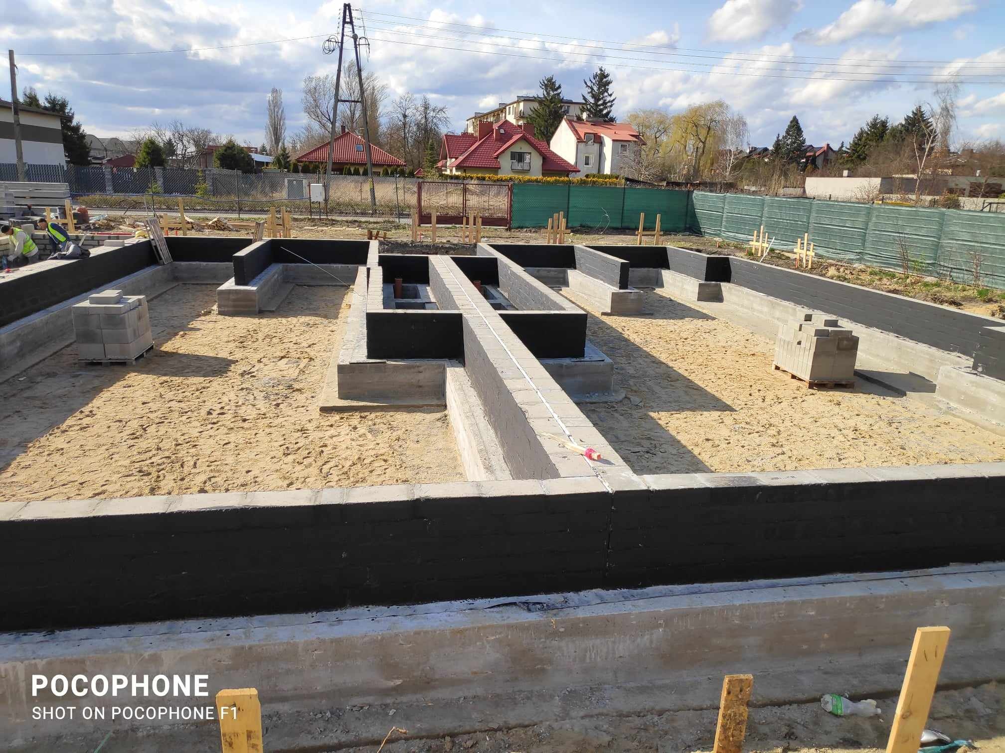 Zalane fundamenty domu z hydroizolacją, widoczne zbrojenia i bloczki betonowe na placu budowy w słoneczny dzień.