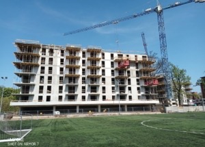 Wielopiętrowy budynek w stanie surowym otwartym, widoczne szalunki stropowe, w tle żurawie budowlane i boisko sportowe.