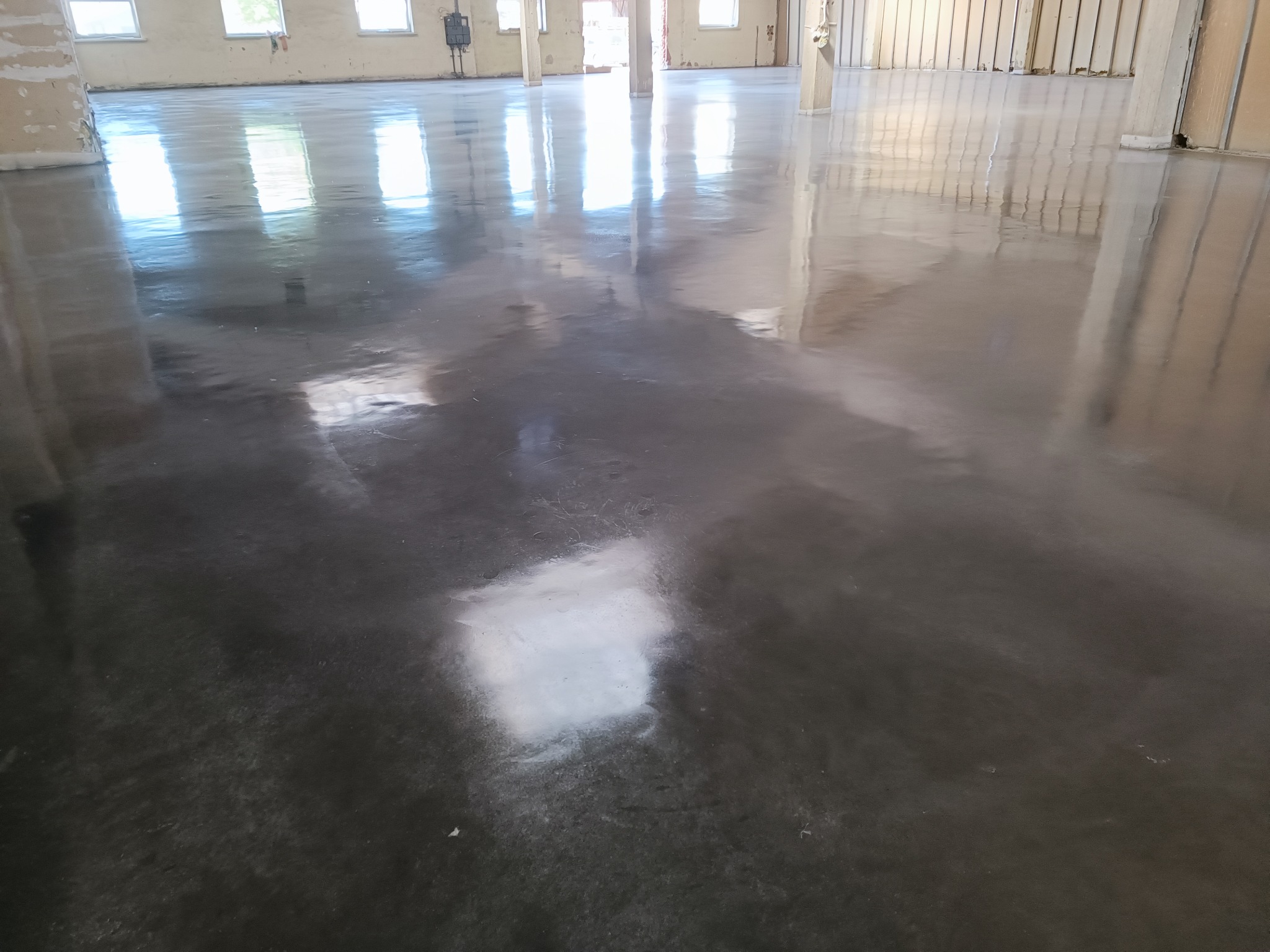 Wnętrze budynku w trakcie remontu z nową, błyszczącą, szarą, betonową posadzką odbijającą światło z okien. Widoczne surowe ściany i filary.