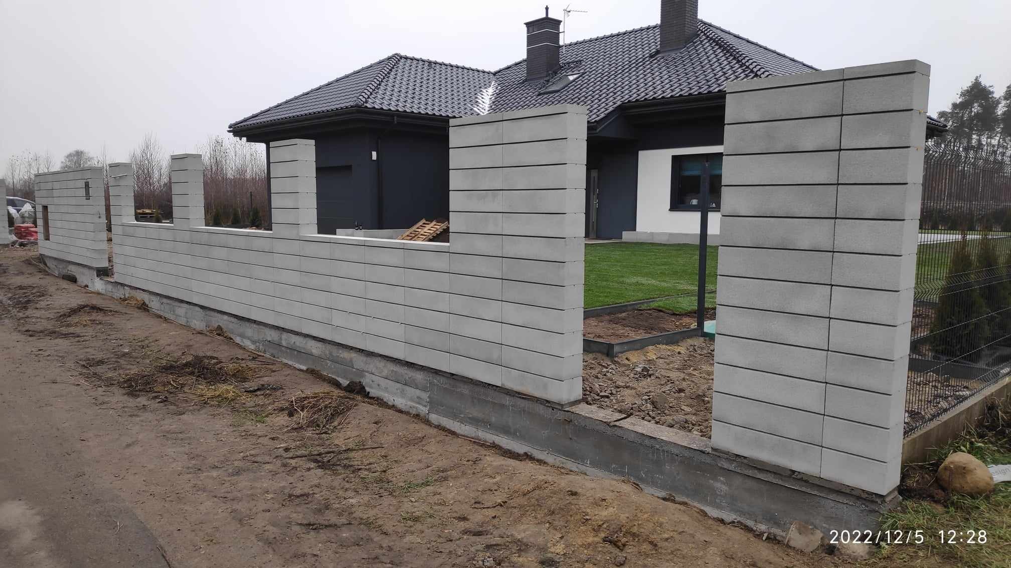 Wznoszenie ogrodzenia z prefabrykowanych betonowych elementów modułowych, widoczny fundament i częściowo ułożona darń na tle nowoczesnego domu z ciemną dachówką.