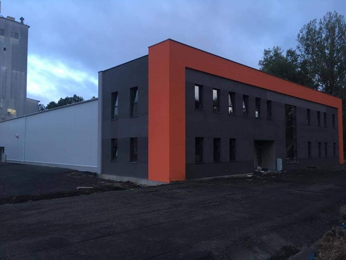 Nowoczesny budynek biurowy z szarą elewacją i pomarańczowymi akcentami, widoczny z zewnątrz na tle pochmurnego nieba.