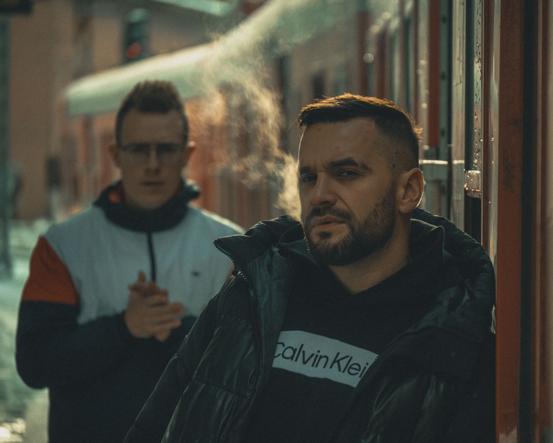 Portret dwóch mężczyzn na tle wagonu kolejowego w zimowej scenerii, główny model w czarnej kurtce z logo, drugi w tle z zamyślonym wyrazem twarzy.