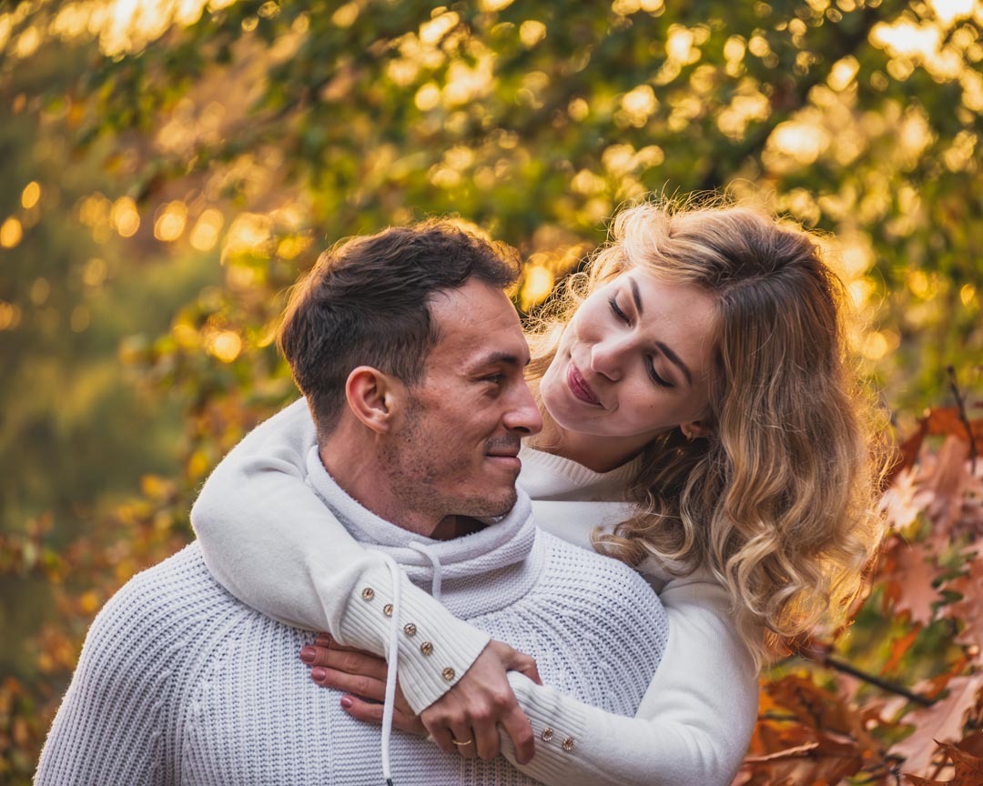 Zakochana para w białych swetrach przytula się na tle jesiennych liści, kobieta z blond włosami spogląda z czułością na mężczyznę.