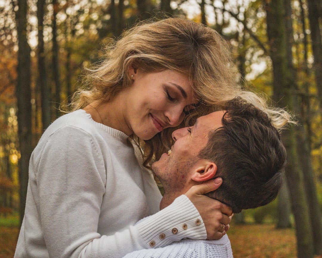 Zakochana para w jesiennym lesie, kobieta obejmuje mężczyznę za szyję, oboje uśmiechnięci, ciepłe światło.