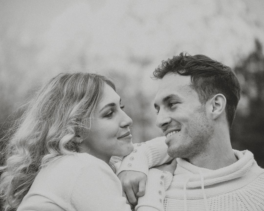 Portret zakochanej pary w swetrach, spoglądających na siebie z uśmiechem, w czarno-białej tonacji.