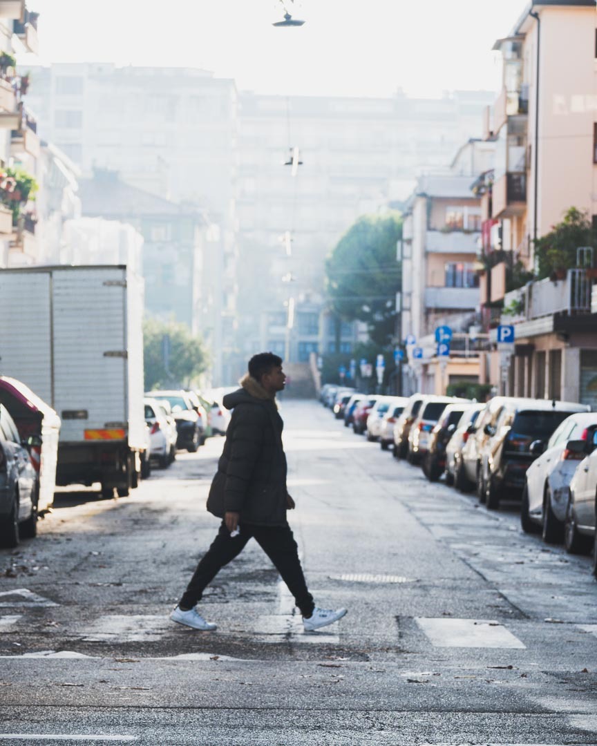 Mężczyzna w zimowej kurtce przechodzi przez pasy na mokrej, miejskiej ulicy, otoczonej rzędami zaparkowanych samochodów i budynkami w tle, w porannym, lekko zamglonym świetle.