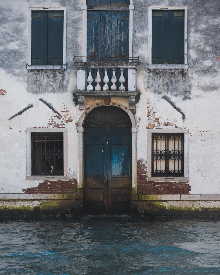 Fasada weneckiego budynku z balkonem i oknami z okiennicami, częściowo zalana wodą, widoczne ślady wilgoci i erozji na murze.