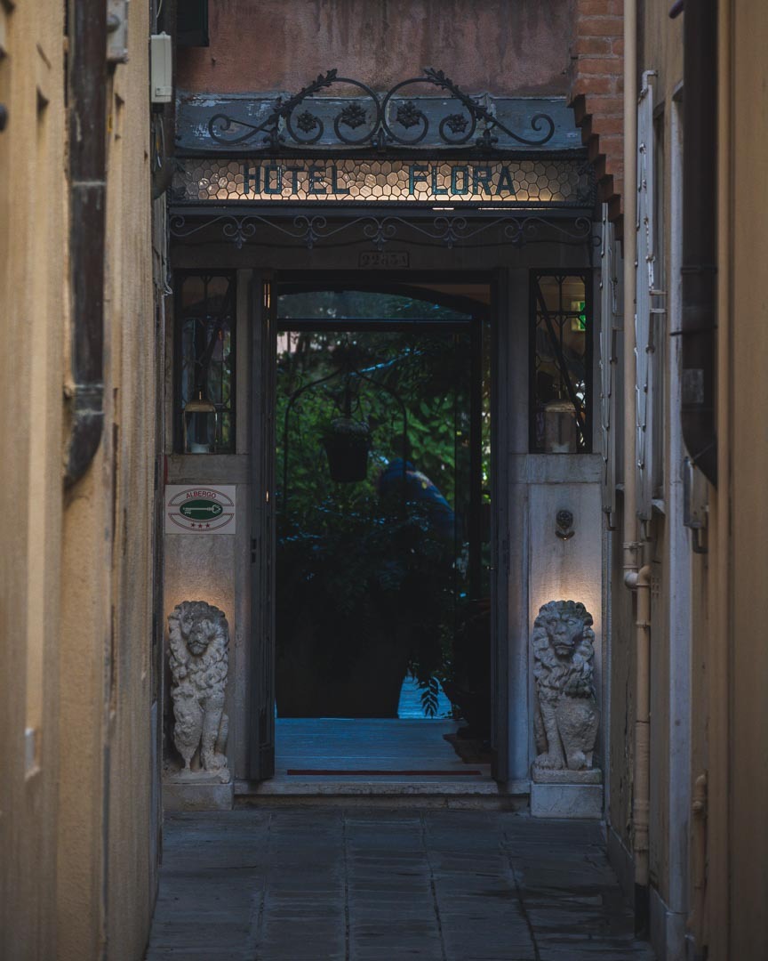 Wąskie przejście do Hotelu Flora w Tarnowskich Górach, z kamiennymi lwami strzegącymi wejścia, oświetlonym szyldem i zielenią widoczną w głębi.