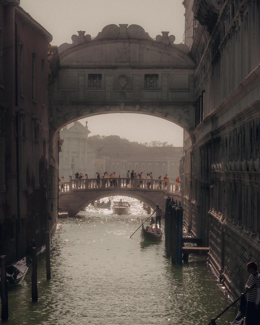 Wenecja: Gondola przepływa pod Mostem Westchnień, widok z perspektywy kanału, refleksy słońca na wodzie.