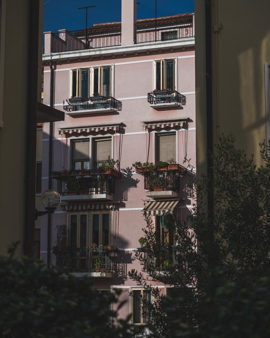 Fasada kamienicy w pastelowym różu z balkonami i roślinami, widziana zza zieleni krzewów, w słoneczny dzień.