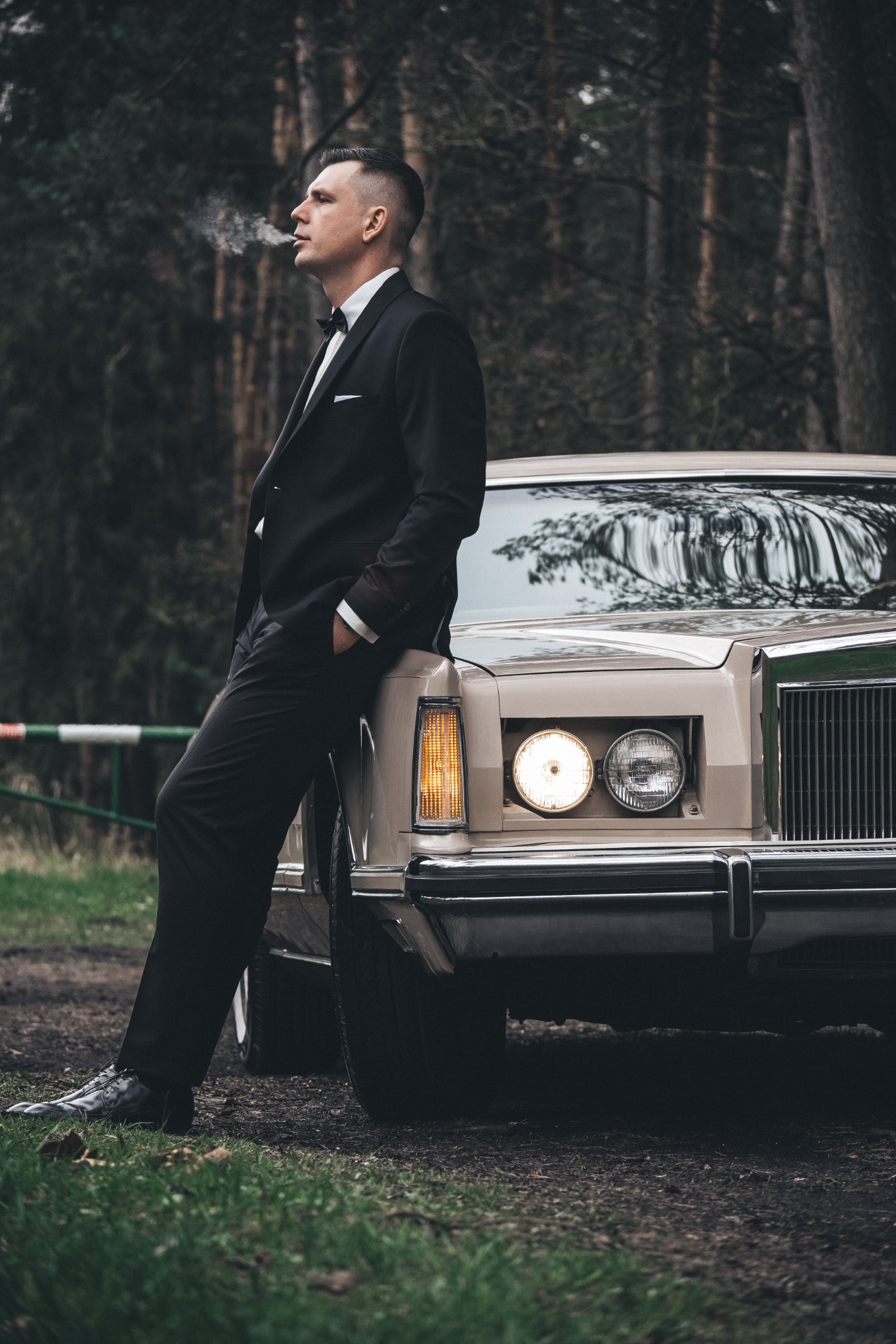 Elegancki mężczyzna w czarnym garniturze i muszce, oparty o beżowy klasyczny samochód w leśnej scenerii, z dymem z papierosa unoszącym się w powietrzu.