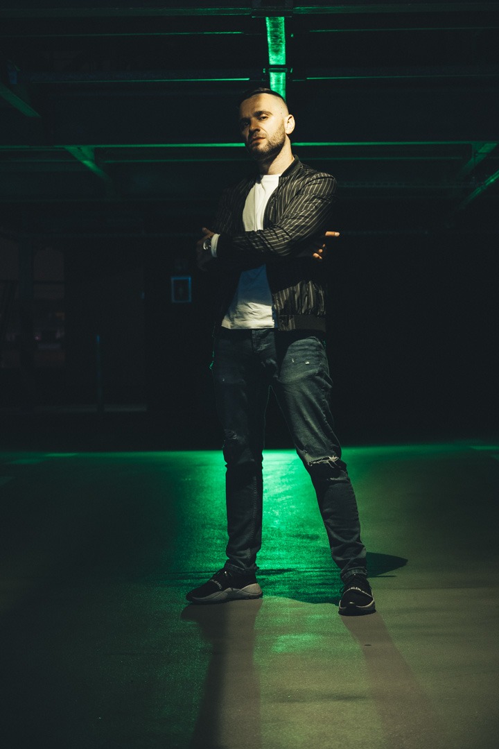 Mężczyzna w jeansach i kurtce w paski, stojący na betonowej podłodze w zielonym oświetleniu, z rękami skrzyżowanymi na piersiach, patrzący wprost w obiektyw.