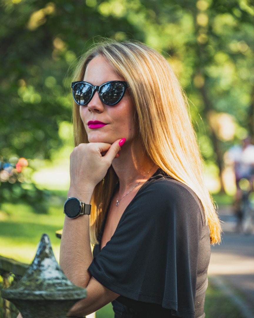 Portret kobiety w czarnych okularach przeciwsłonecznych, z ręką podpartą pod brodą, różowe usta i paznokcie, na zewnątrz w parku z zamazanym tłem drzew i ścieżki.