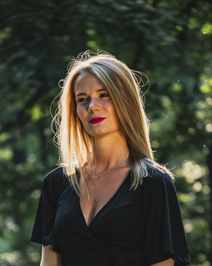 Portret blondynki w czarnej sukience na tle rozmytej zieleni, z promieniami słońca oświetlającymi włosy i twarz, subtelna biżuteria na szyi.