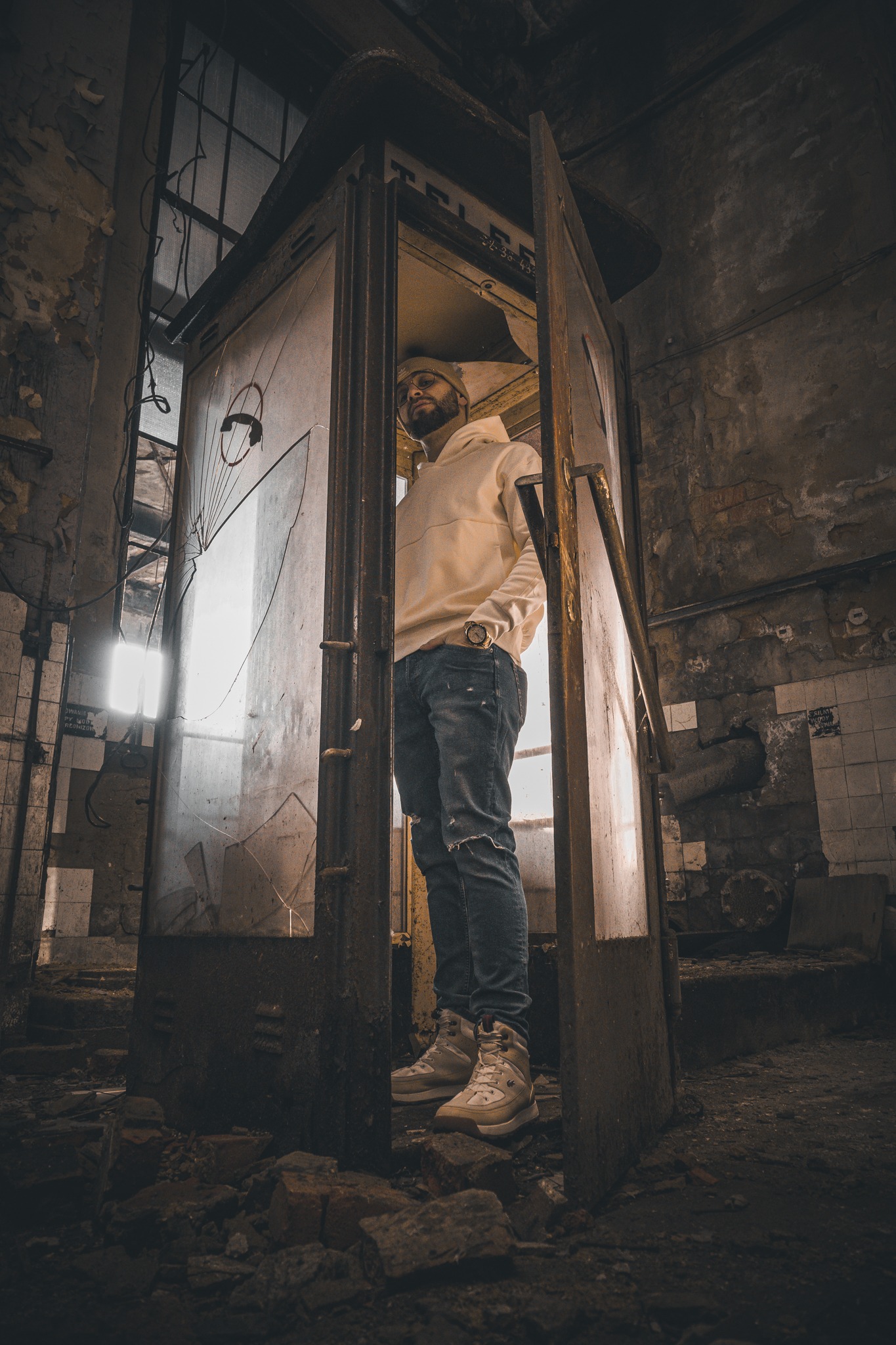 Mężczyzna w beżowej bluzie z kapturem i dżinsach pozuje w zniszczonej budce telefonicznej z powybijanymi szybami, w opuszczonym, industrialnym wnętrzu.