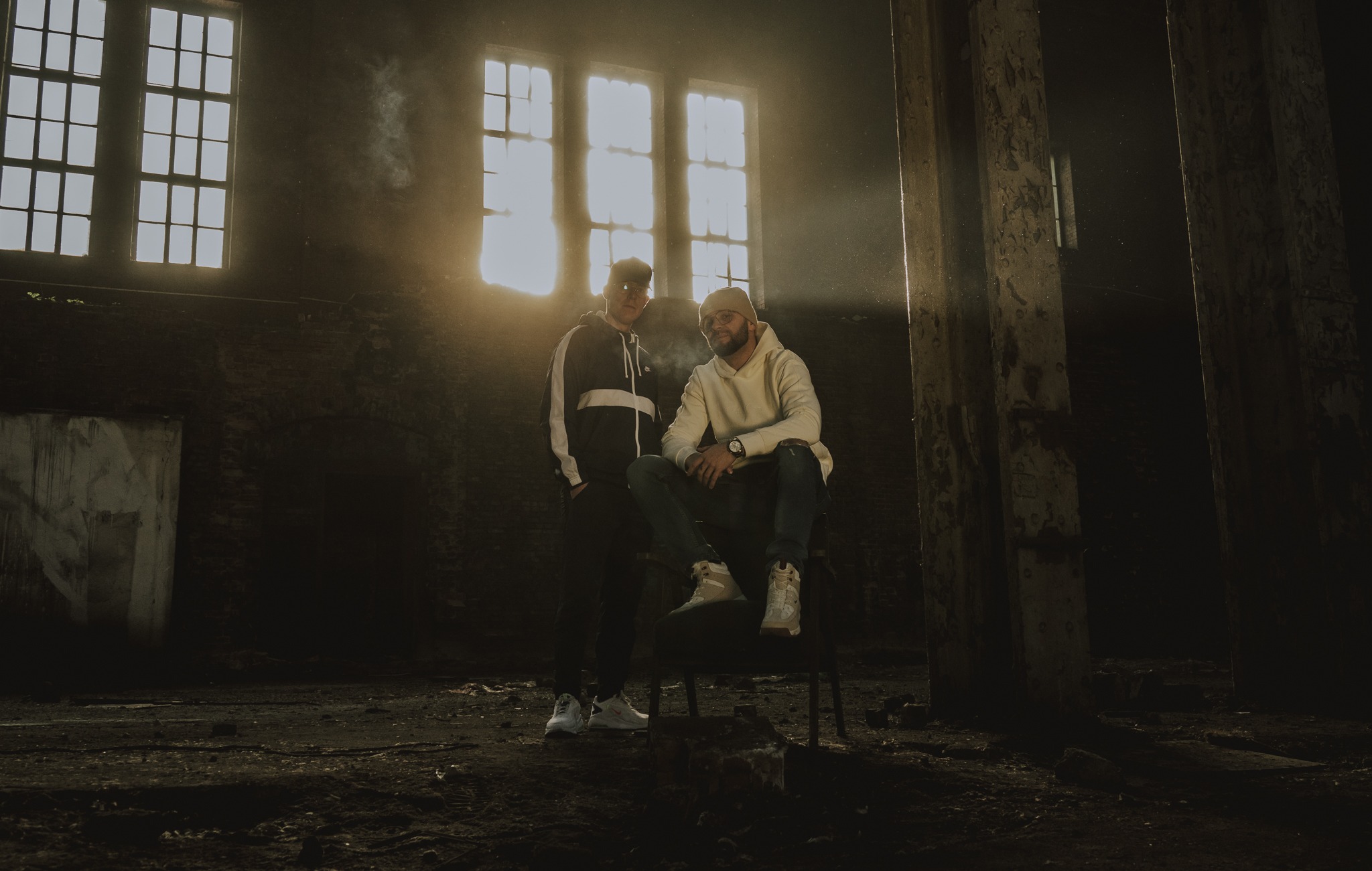 Dwóch mężczyzn w sportowych strojach pozuje w opuszczonym, industrialnym wnętrzu z promieniami światła wpadającymi przez okna, tworząc dramatyczny efekt.