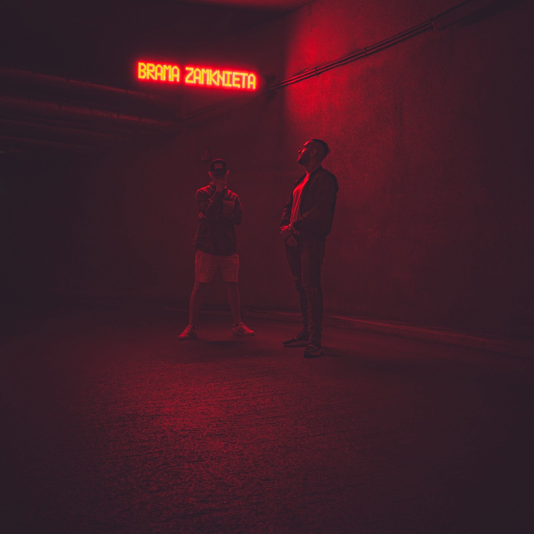 Dwaj mężczyźni stojący w ciemnym tunelu oświetlonym czerwonym neonem z napisem 'Brama Zamknięta'.