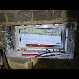 Michał Stąpor - Montaż okna PCV w surowym murze z widocznymi poziomnicami, klinami i fragmentami narzędzi, ukazujący etap prac wykończeniowych.