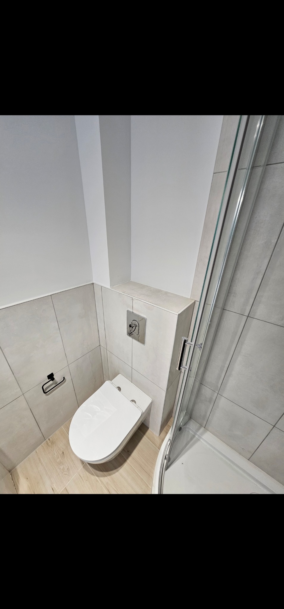 Nowoczesna łazienka z podwieszaną toaletą, szarą glazurą i kabiną prysznicową z chromowanymi detalami, drewnopodobna podłoga.