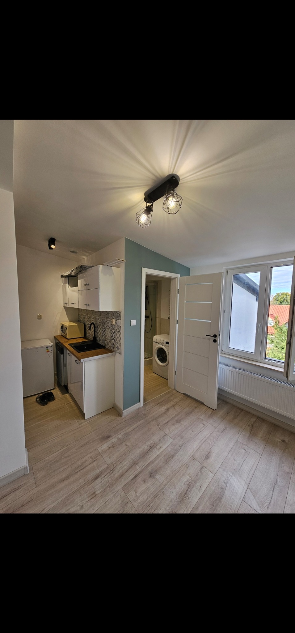 Widok na odnowione wnętrze mieszkania z nową podłogą z paneli, aneksem kuchennym z białymi szafkami i czarnym zlewem, oraz łazienką z pralką widoczną w tle.