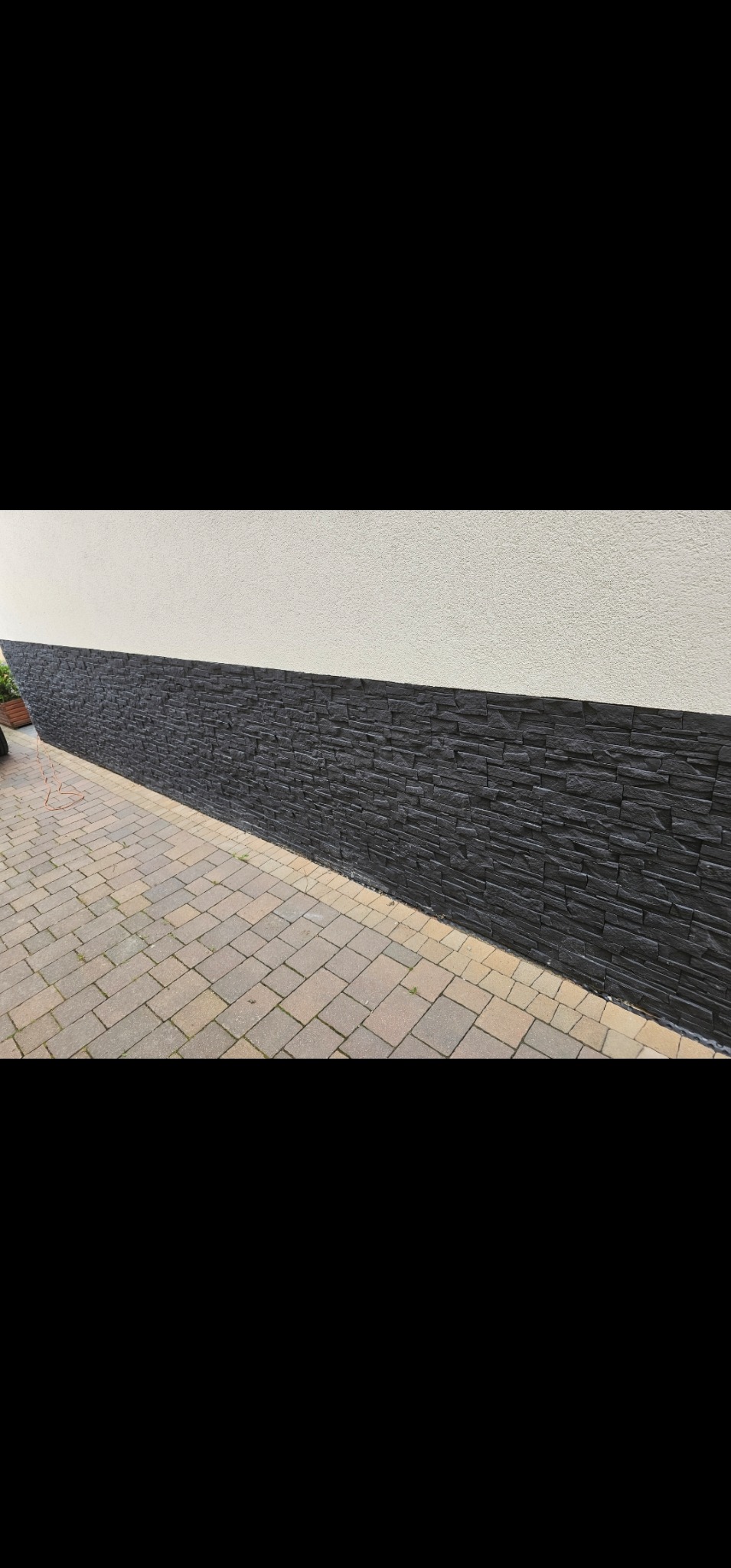 Część elewacji budynku z wykończeniem w postaci czarnej, kamiennej okładziny na dole i białym tynkiem powyżej, widoczne również fragmenty chodnika z kostki brukowej.