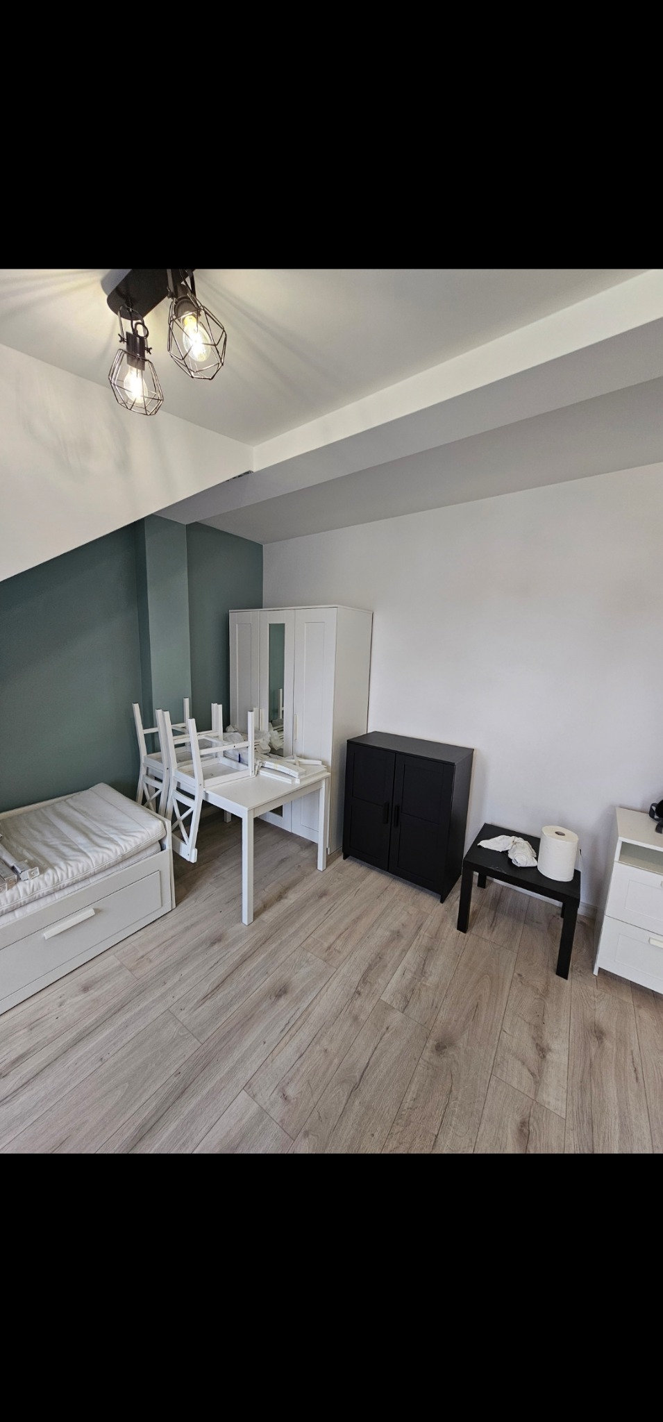 Pusta przestrzeń w jasnych kolorach z podłogą imitującą drewno, białymi meblami i ścianą w odcieniu zieleni, oświetlona przez lampę sufitową z żarówkami w stylu retro.