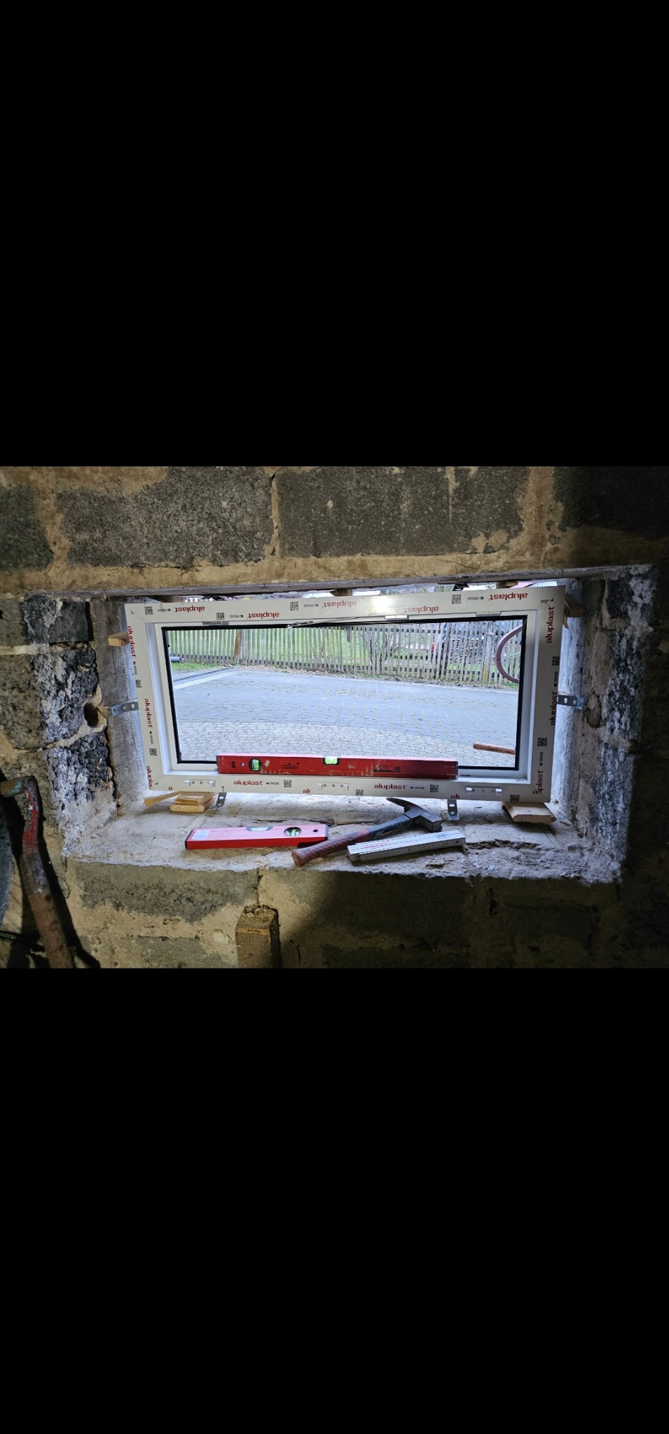 Montaż okna PCV w surowym murze z widocznymi poziomnicami, klinami i fragmentami narzędzi, ukazujący etap prac wykończeniowych.