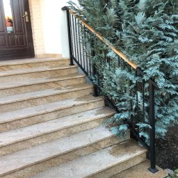 B&B FUTURE Krystian Bober - Zewnętrzne schody z beżowego kamienia z czarną metalową balustradą i drewnianym poręczą, obok rośnie ozdobne drzewko iglaste.