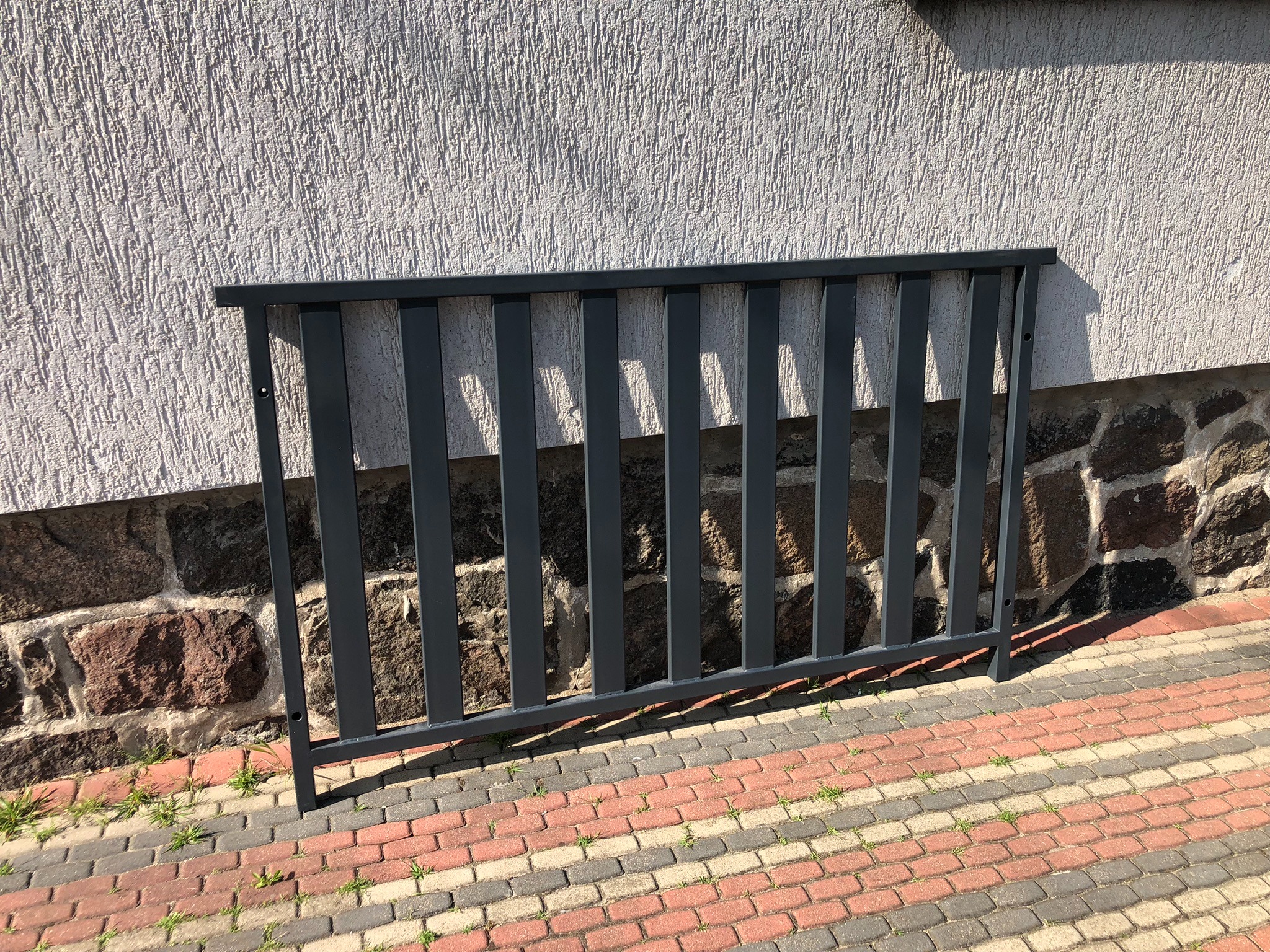 Ciemnoszara metalowa konstrukcja z pionowych prętów, prawdopodobnie fragment ogrodzenia, oparta o ścianę budynku z tynkiem strukturalnym i kamienną podmurówką, na pierwszym planie kostka brukowa.