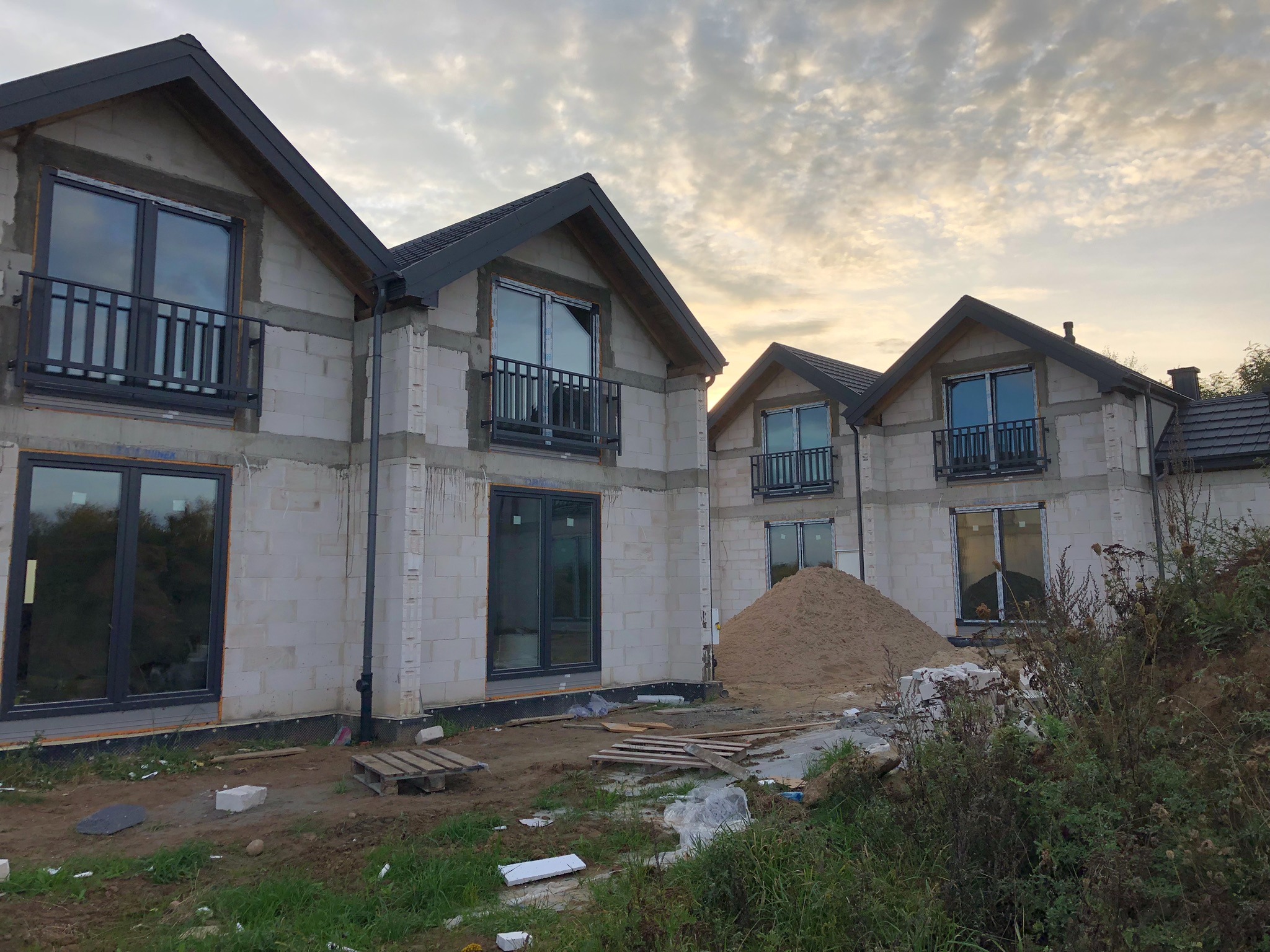 Surowe budynki w Nidzicy z czarnymi balustradami balkonowymi i stertą piasku na placu budowy, uchwycone o zmierzchu.