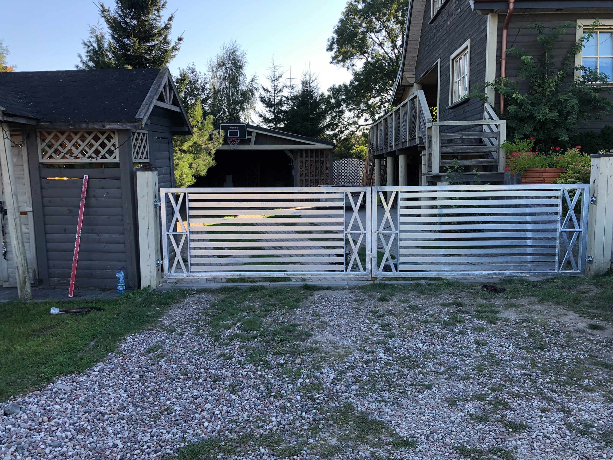 Nowoczesna, metalowa brama wjazdowa z poziomymi szczeblami i dekoracyjnymi krzyżami, prowadząca na żwirowy podjazd przed domem z ciemnego drewna.