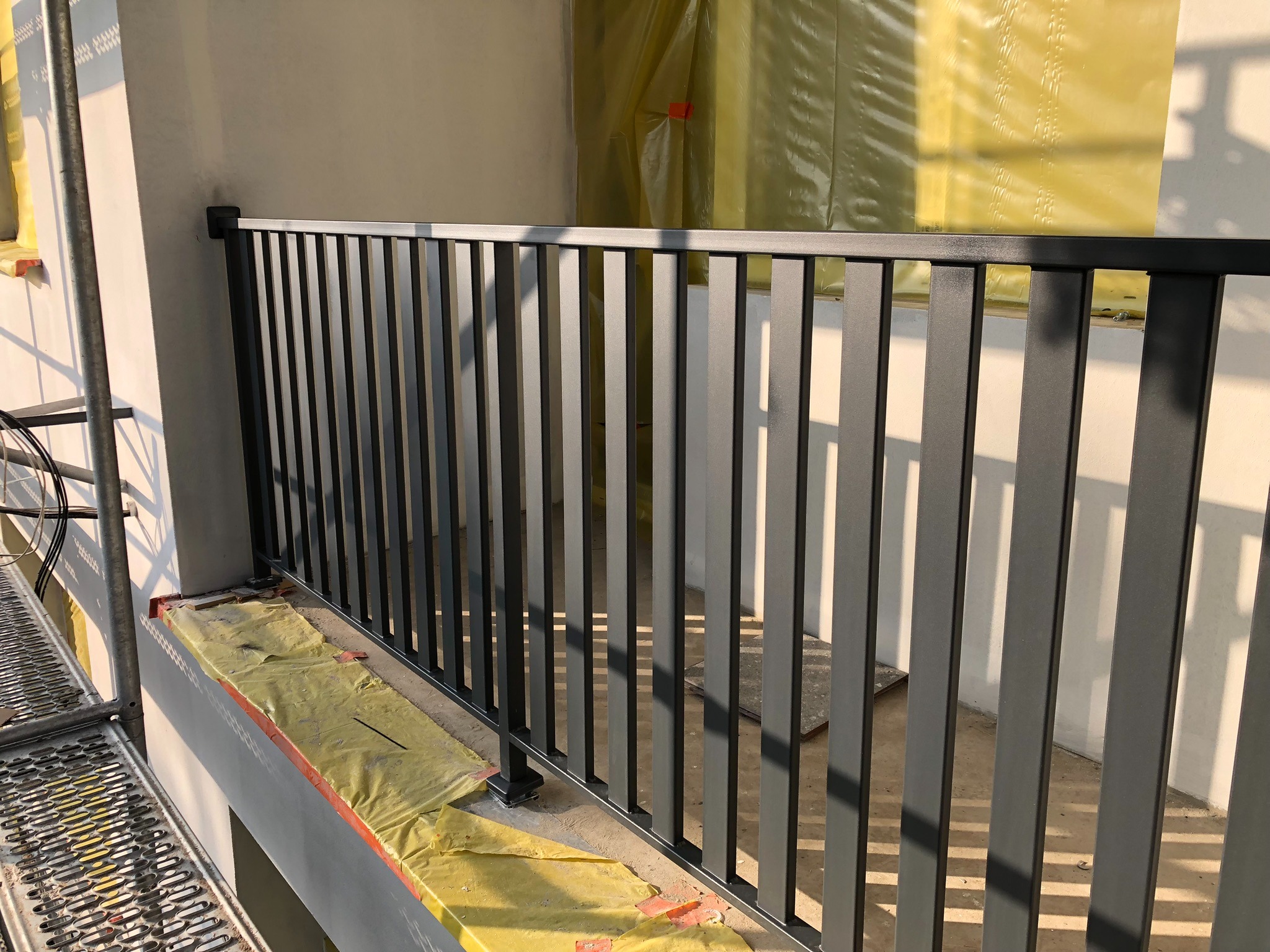 Nowoczesna, grafitowa balustrada balkonowa z pionowych profili, zabezpieczona folią malarską, widoczna ze strony rusztowania.