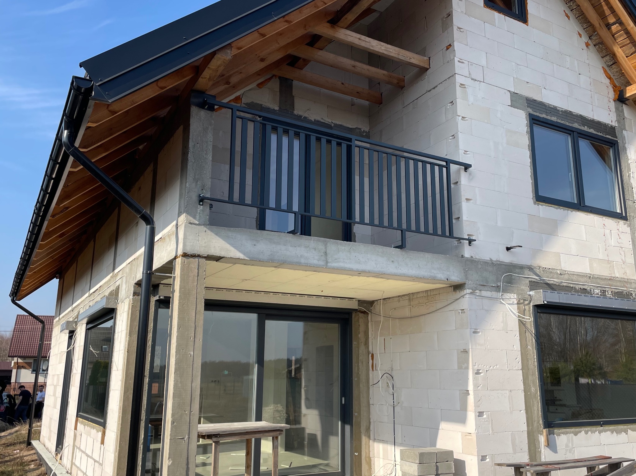 Surowy stan budowy domu z widoczną szarą, metalową balustradą balkonową, czarnymi ramami okiennymi i rynnami, ściany z białego betonu komórkowego, drewniana konstrukcja dachu w trakcie montażu.