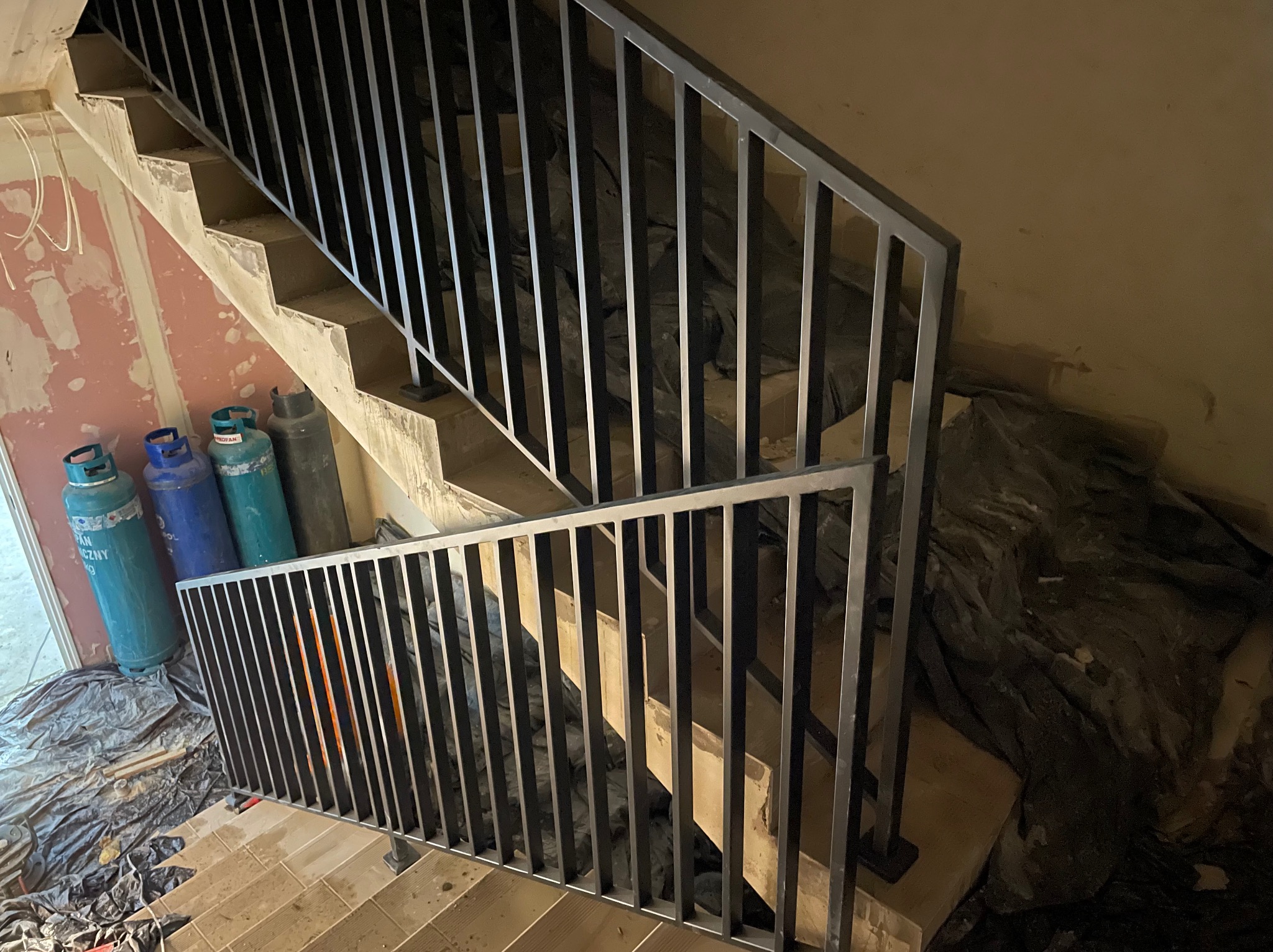 Surowe schody z metalową balustradą w trakcie montażu; widoczne butle gazowe i folia ochronna.