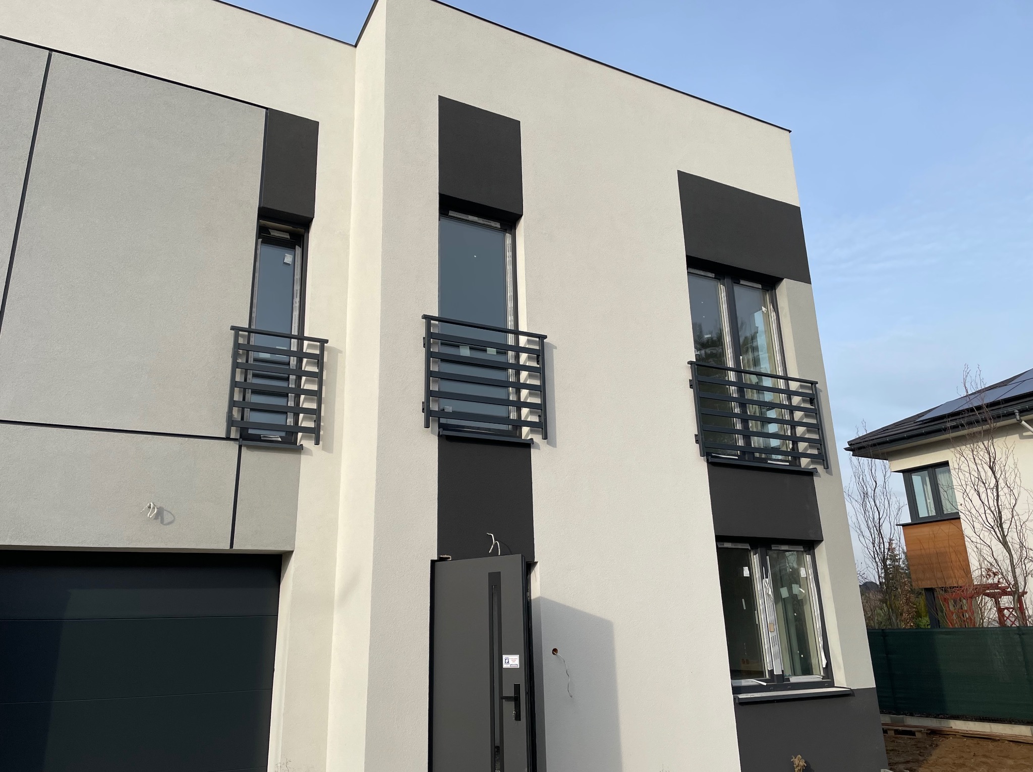 Nowoczesny dom z szarą elewacją, czarnymi akcentami i minimalistycznymi balustradami okiennymi w kształcie poziomych prętów.
