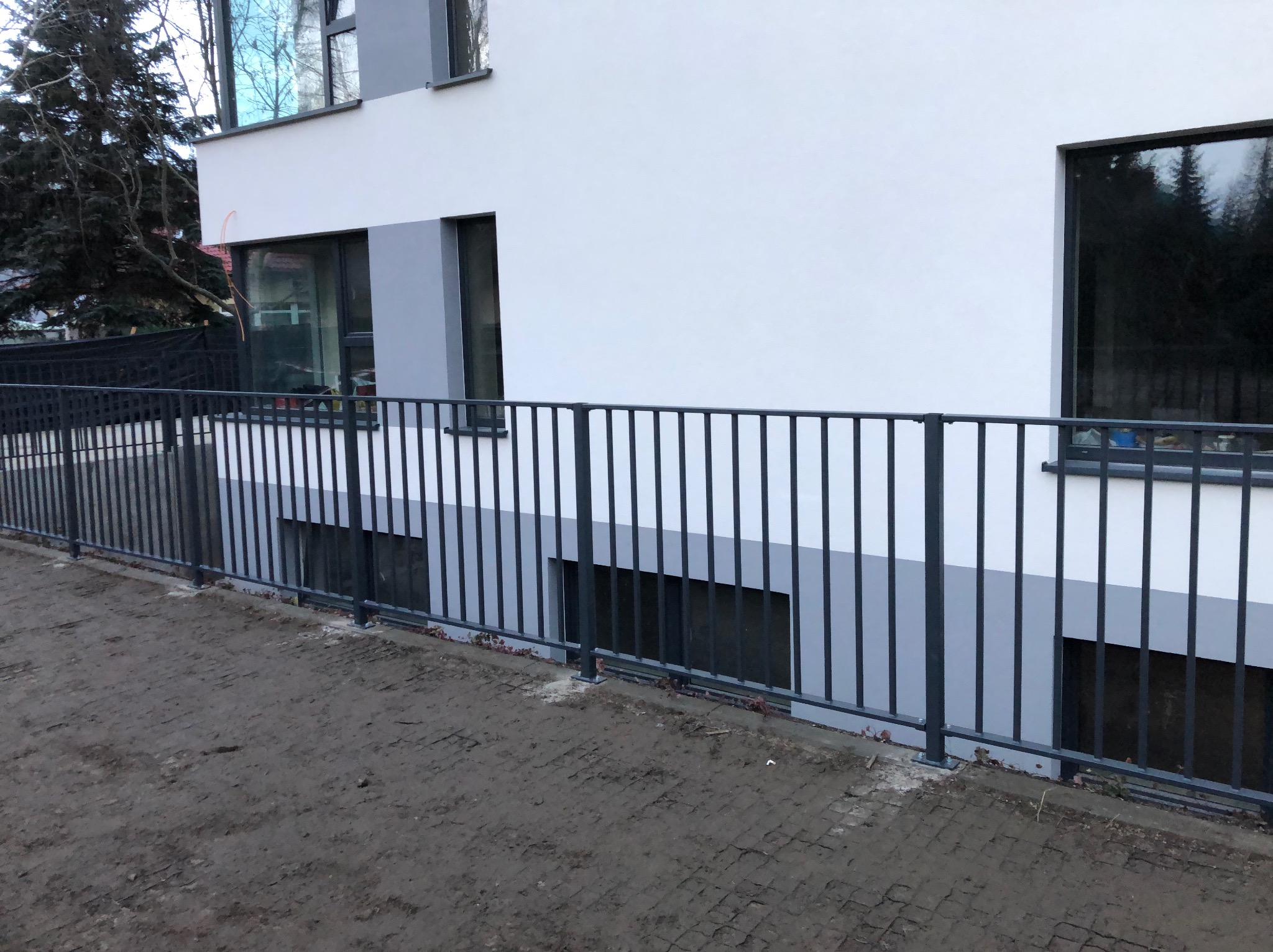 Nowoczesne ogrodzenie metalowe w kolorze grafitowym, otaczające budynek z szarymi oknami, widoczne ślady błota na ziemi.