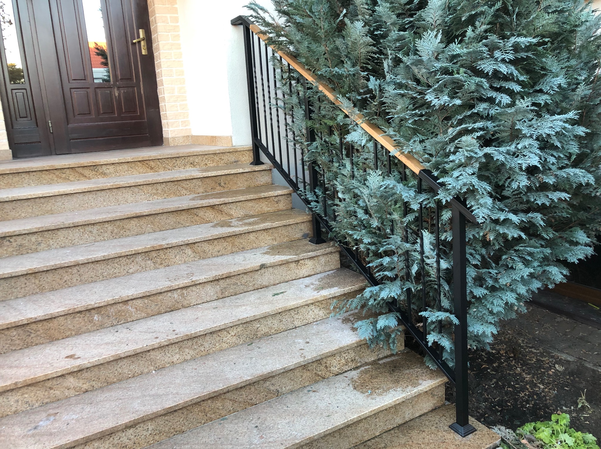 Zewnętrzne schody z beżowego kamienia z czarną metalową balustradą i drewnianym poręczą, obok rośnie ozdobne drzewko iglaste.
