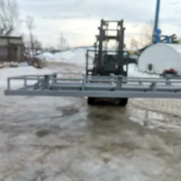 Wózek widłowy przewożący metalową konstrukcję na zewnątrz, w tle budynek i pozostałości śniegu.