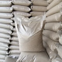 Stos worków z beżowym kruszywem, jeden worek przezroczysty, ukazujący zawartość, ułożone równo jeden na drugim pod ścianą.