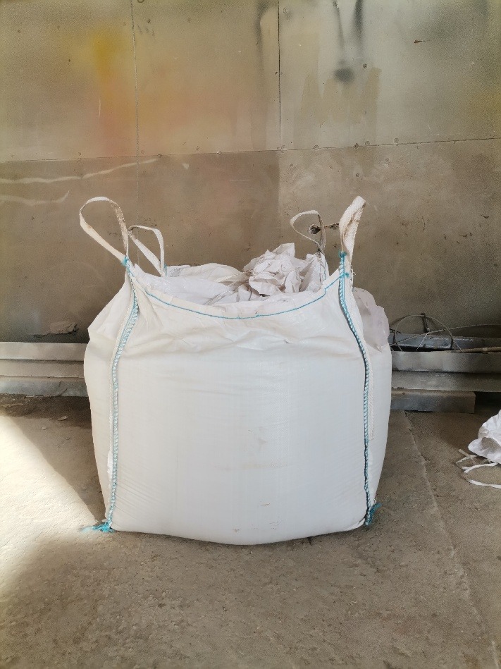 Worek typu Big Bag wypełniony materiałem, stojący w pomieszczeniu o szarych ścianach.