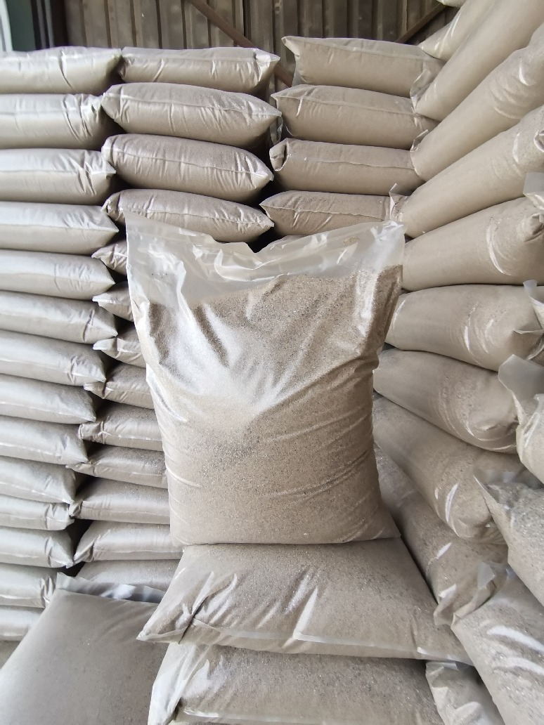 Stos worków z beżowym kruszywem, jeden worek przezroczysty, ukazujący zawartość, ułożone równo jeden na drugim pod ścianą.