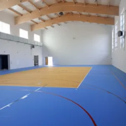 Sala gimnastyczna z nową, niebiesko-żółtą nawierzchnią sportową z widocznymi liniami boisk, drewnianą konstrukcją dachu i białymi ścianami.