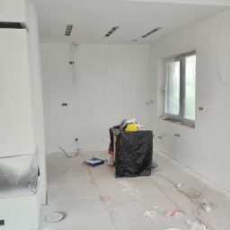 Trzy kolory - Wnętrze pokoju po malowaniu na biało, widoczne zabezpieczone okno folią, zakryte meble, narzędzia malarskie i ślady farby na podłodze.