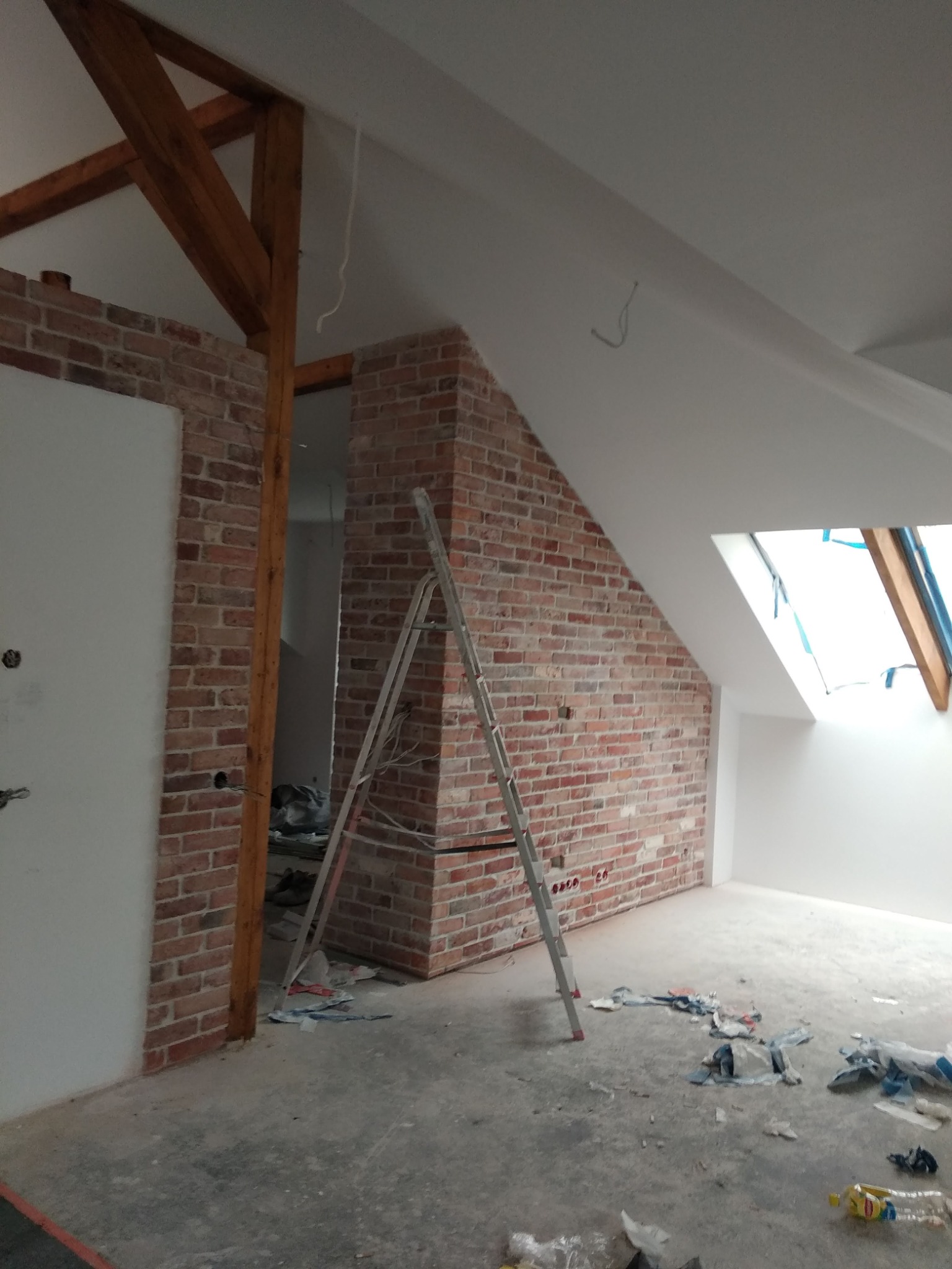 Poddasze w trakcie wykańczania, ceglana ściana z drewnianą belką, biała ściana i sufit, aluminiowa drabina oparta o ścianę, widoczne ślady prac remontowych na podłodze.