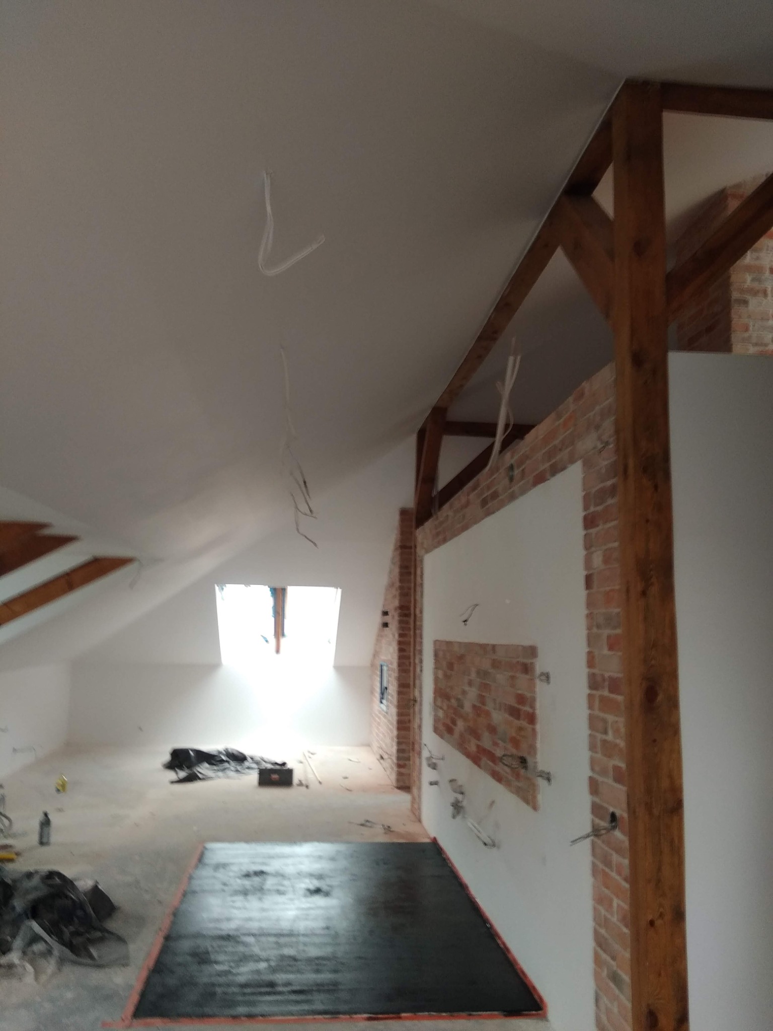Poddasze w trakcie remontu: świeżo pomalowane ściany na biało, fragment ściany z cegły, drewniane belki konstrukcyjne i kable zwisające z sufitu.