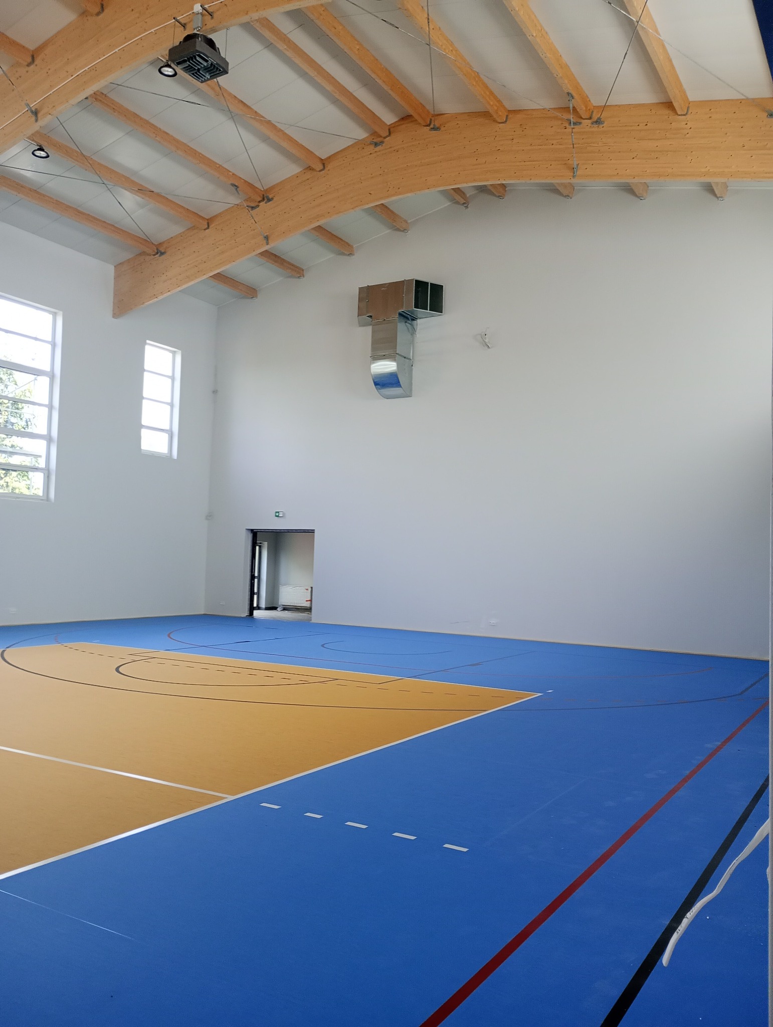 Nowo wykończona hala sportowa z parkietem w kolorach niebieskim i żółtym, drewnianą konstrukcją dachu i białymi ścianami, widoczne okna i system wentylacji.