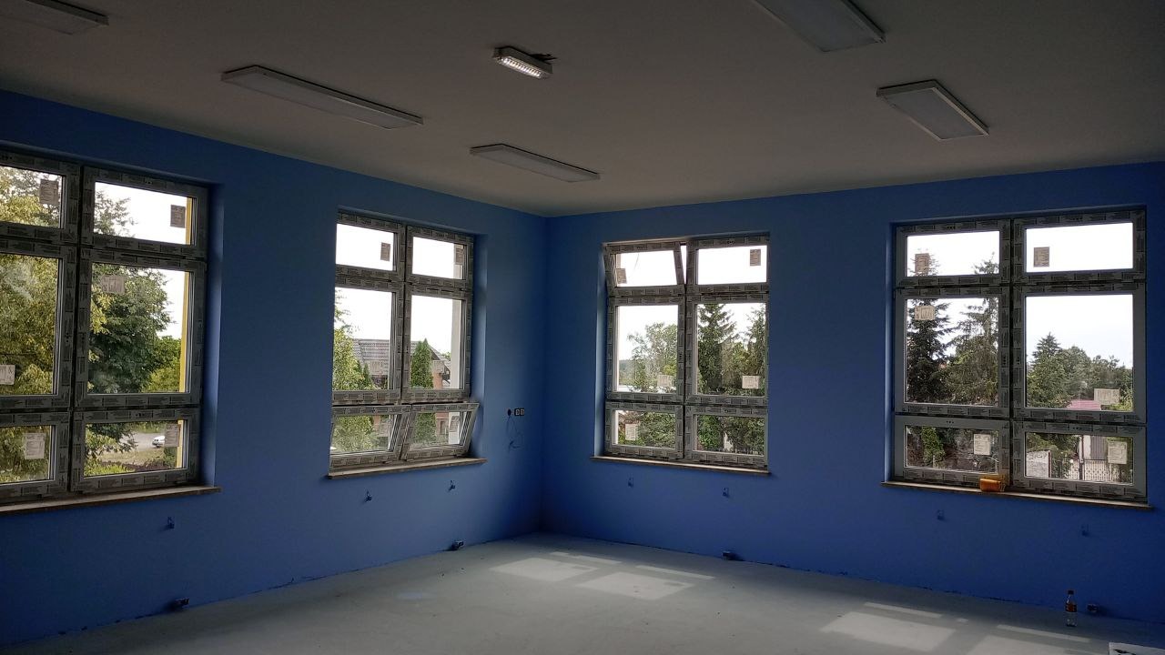 Jasne, błękitne wnętrze z czterema oknami, widok na zewnątrz z zielenią drzew, surowe wykończenie podłogi, widoczne punkty świetlne na suficie, ściany świeżo pomalowane.