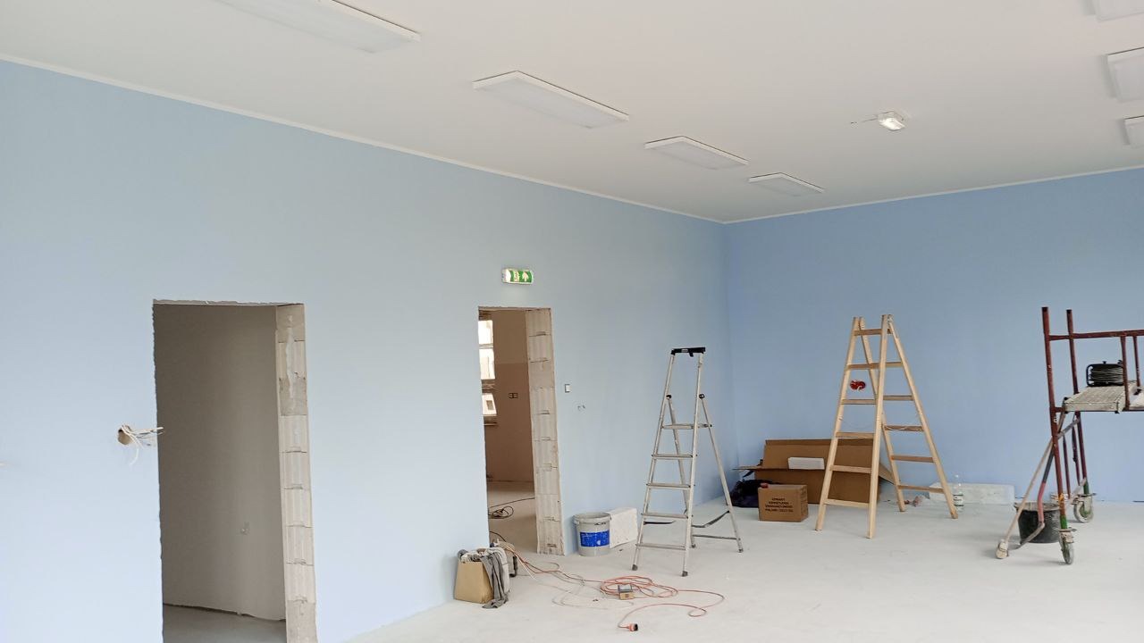 Świeżo pomalowane na błękitno ściany w biurze, widoczne drabiny malarskie, sufit z lampami kasetonowymi, przygotowane kable i puszka z farbą.